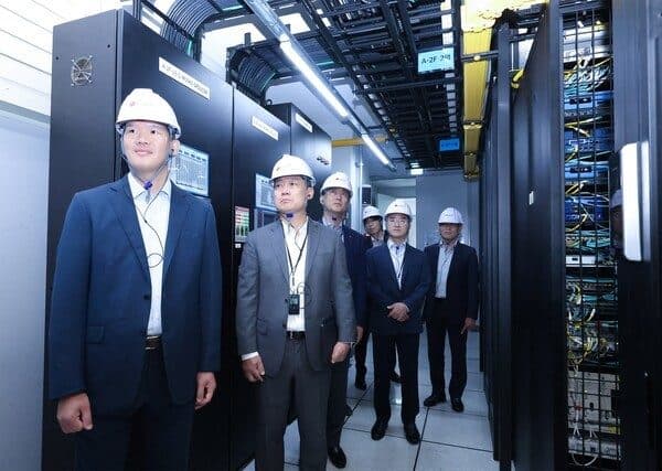 Executives in business suits and white hard hats walking down an aisle of black server racks and overhead cable trays during a data-centre walkthrough.